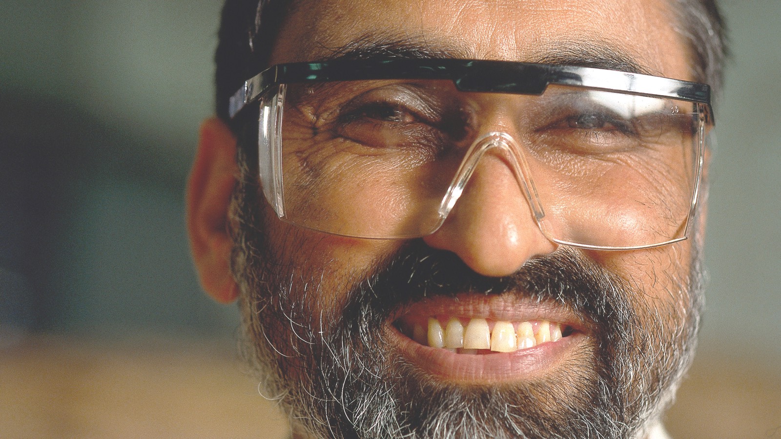 A bearded man wearing safety glasses stands in a factory and smiles.