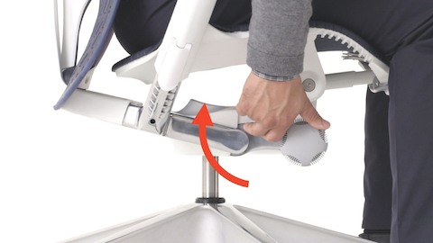 Una vista baja de una silla Mirra 2 con marco blanco cuando una persona se sienta en ella y demuestra el ajuste de la perilla de control de tensión de inclinación.
