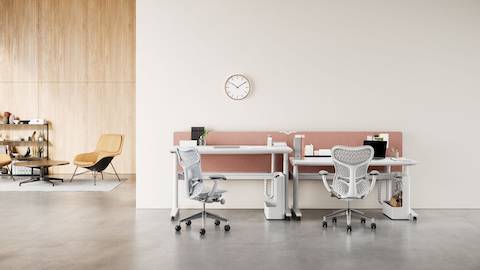 An office setting with a pair of Augment Ratio desks in the foreground with white worksurfaces and grey Mirra 2 Chairs. A lounge area with Striad Lounge Chairs, a Civic Table, and a bookcase in the background. One of the desks is raised to standing height.