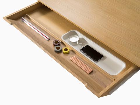 The main drawer of a light wood Carafe Table, open to reveal a ruler, tape, pencils, and smartphone inside.