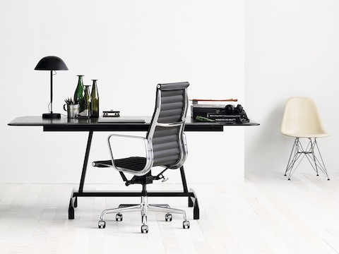 Pequeña oficina con una silla Eames Aluminum Group negra, una mesa AGL negra y una silla de fibra de vidrio moldeada Eames de color blanco.