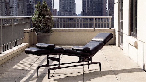 Un cuero negro Eames Chaise situado en un balcón con vistas a un horizonte urbano.