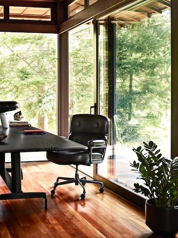 Una silla ejecutiva Eames de cuero negro con una mesa AGL negra en una oficina hogareña con paredes de vidrio con vistas a los árboles.