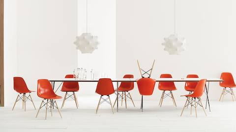 Un esquema de sillas de visita de fibra de vidrio moldeada Eames en rojo anaranjado con patas espigadas en arce y mesa Nelson con patas en X.