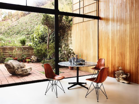 Tres sillas laterales de madera moldeada Eames y una mesa redonda de Eames en una habitación con paredes de vidrio con vistas al jardín.