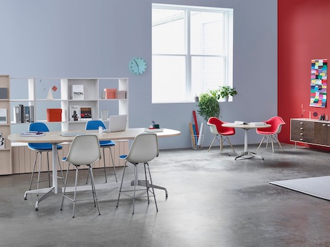 Mesas Eames ovaladas y redondas en un área de colaboración que también cuenta con sillones y taburetes de plástico moldeado Eames en azul y rojo.