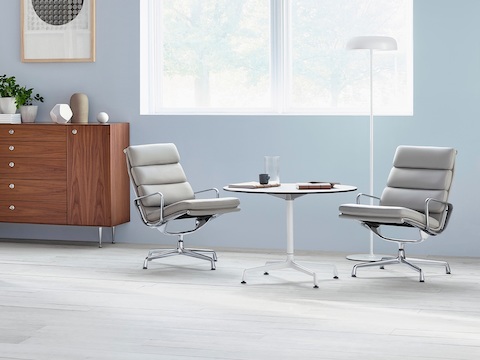 Una mesa redonda de Eames con una parte superior blanca, combinada con dos sillas de Eames Soft Pad de color gris claro.
