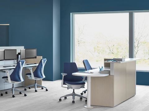 An Ethospace caregiver station in a light wood finish, consisting of a reception desk and a separate workstation with blue upholstered Sayl chairs.
