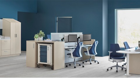 An Ethospace nurse station in a light wood finish, consisting of a reception desk, two-sided workstation for sitting or standing, Mora casework in matching wood, and a supply cart with blue drawers.