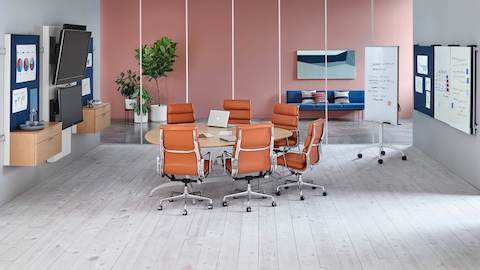 Seis Eames Soft Pad Chairs de color naranja quemado rodean una mesa en forma de lágrima en un espacio de colaboración con elementos de visualización Exclave.