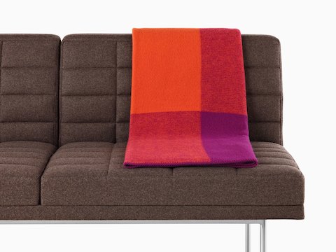 A Girard Throw blanket in shades of orange and magenta folded over the back of a brown Tuxedo Sofa.