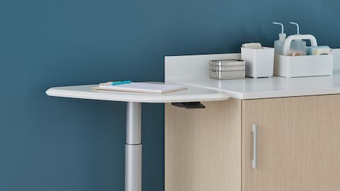 An Intent Solution height-adjustable mobile technology table with a white surface and silver base next to Mora System casework in a light wood finish and Corian surface.