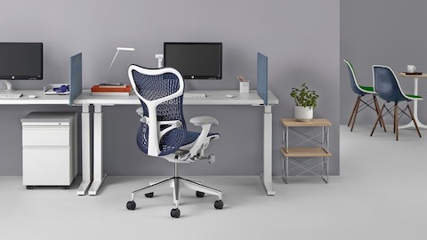 A blue Mirra 2 office chair at a workpoint with a sit-to-stand desk. Select to review Herman Miller's ergonomics research.
