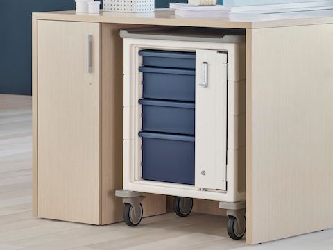 A soft white Procedure and Supply Cart with keyless lock and blue drawers in a storage cove of Ethospace System within a nurses station.