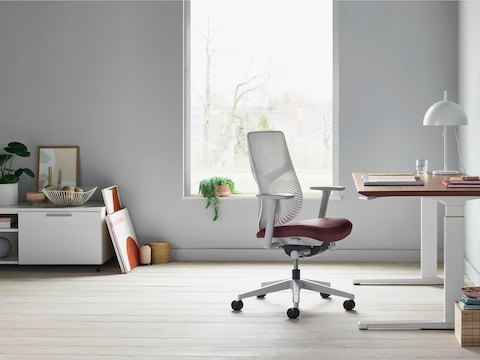 A white Verus Chair with dark red seat next to a Renew Sit-to-Stand Table with a brown surface close to a window.