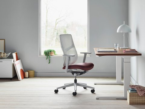 A Verus Chair with a dark red upholstered seat and white Triflex back next to a Renew Sit-to-Stand Table in a home office with gray walls and light hardwood floors.
