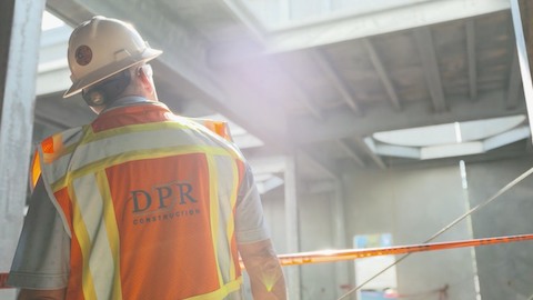 A man, viewed from behind, wears a hard hat and orange safety vest. Select to go to a case study featuring DPR Construction.