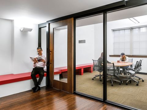 A man reads a textbook while waiting outside of a glass-walled meeting room. 