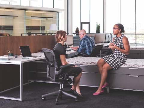 Employees work and converse while seated in an open office environment. 