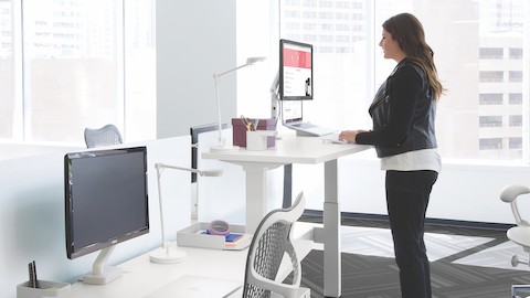 A woman types on a laptop at a standing desk near a window.