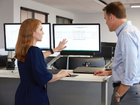 Two people talk while looking at a monitor on a standing desk.