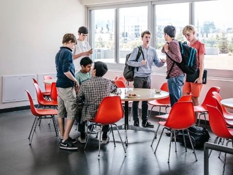 A group of students gather around a space full of tables and Eames molded chairs. 