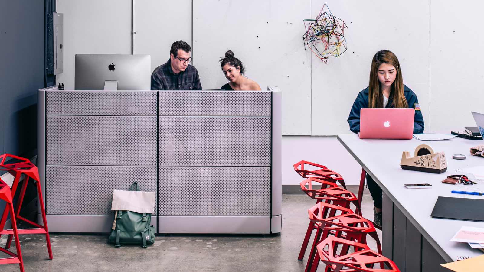 A man and woman sit behind a tall gray workstation divider with a large Apple desktop display. A female student sits nearby atop a red Magis Stool_One at a tall desk with a laptop and various office supplies. 