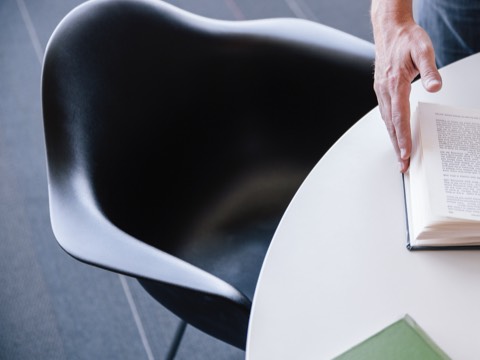 Hands flip the pages of a book that rests on a table served by a black Eames Molded Plastic Chair.