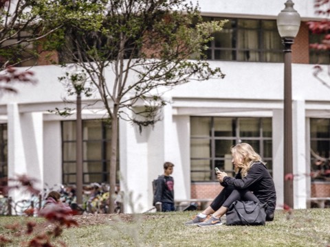 A female college student checks her smartphone while sitting on the grass outside an academic building. 