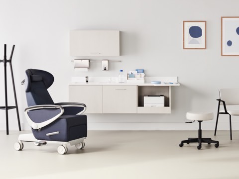 Exam room with a dark blue Ava Recliner and an Intent Solution mobile, height-adjustable table and wall unit to the side with a Physician Stool and Verus Side Chair in a light gray upholstery and black frame with Mora casework in a light wood laminate on the wall in the background.