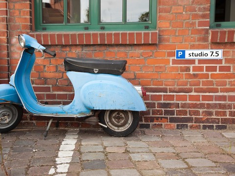 A vintage blue motorbike parked alongside the exterior brick wall of Studio 7.5 in Berlin.