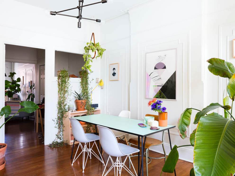 A residential dining area featuring a rectangular table, white Eames Molded Plastic Chairs, modern artwork, and plants.