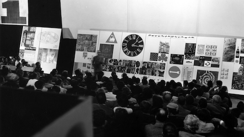 A man speaks before a seated crowd. Select to go to an article about a 1952 lecture by George Nelson, Charles Eames, and Alexander Girard.