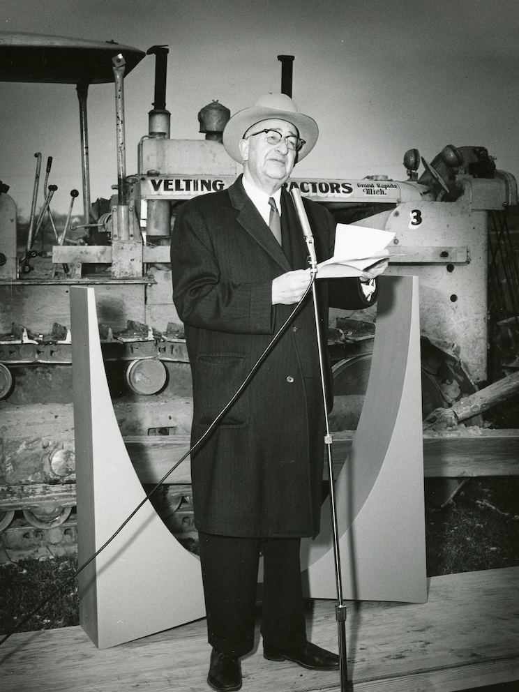 Herman Miller founder D.J. De Pree speaks into a microphone and stands before heavy machinery at a building site.