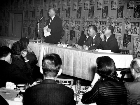A black-and-white photo of Herman Miller founder D.J. De Pree standing behind a head table and speaking at a professional gathering.