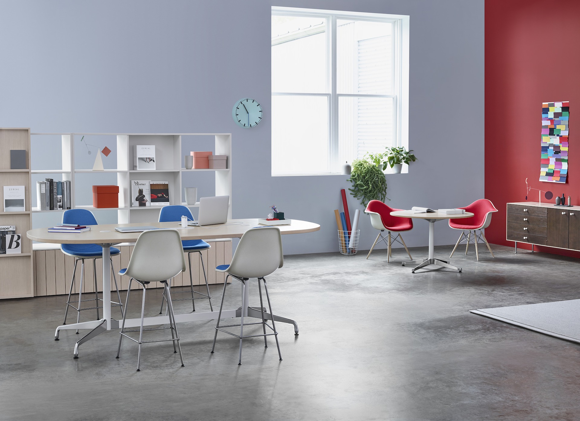 A Workshop setting is built around a group of four Eames Stools around a standing-height Eames Table, with a pair of upholstered Eames Molded Fiberglass armchairs at a small Eames Table below a nearby window.