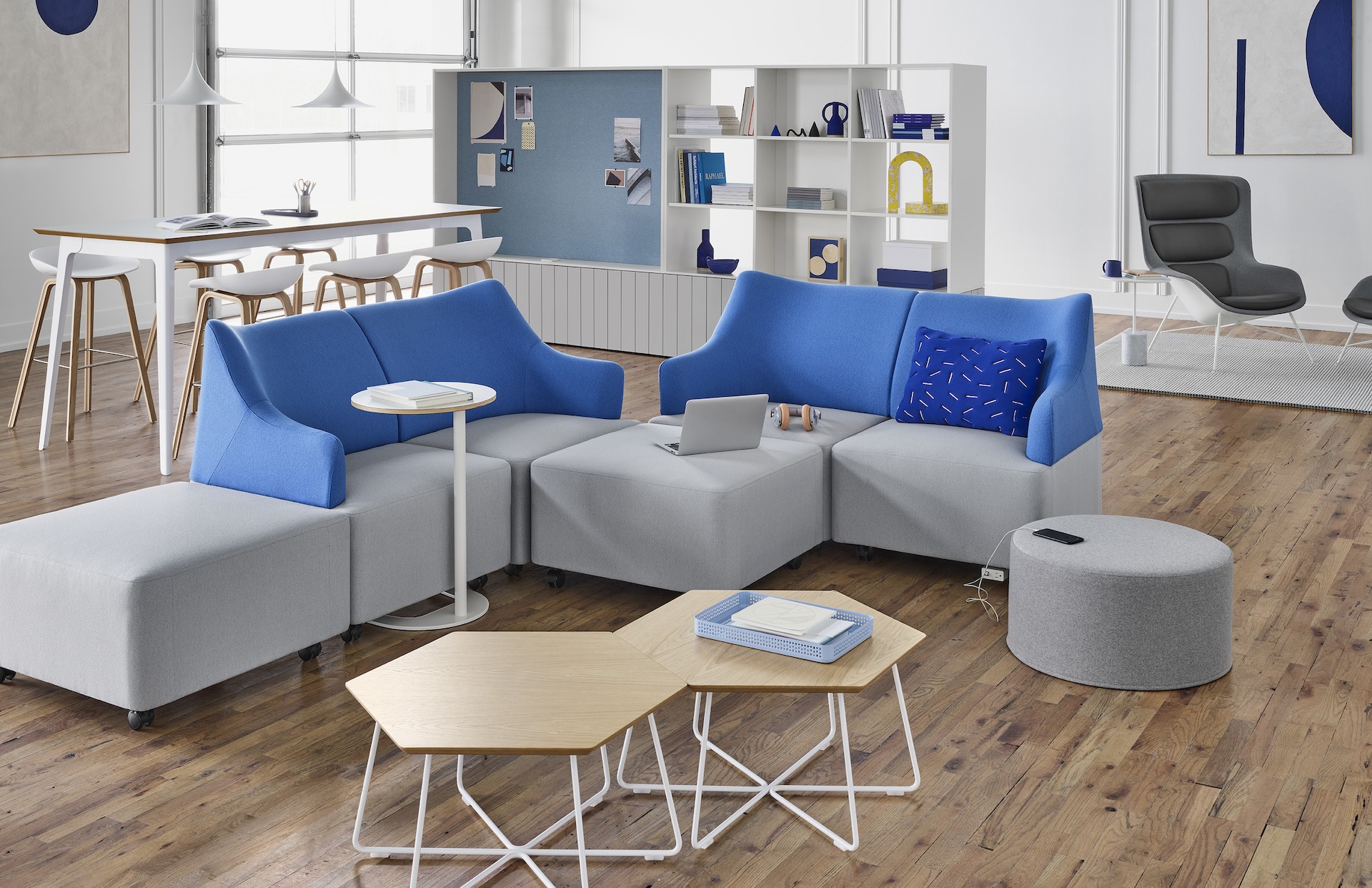 Plex Lounge Furniture in light grey and blue around two Pollen Tables in oak with white bases, a light grey wide Drum Pouf, and a white Plex Side Table. The scene is set in front of a work space and another lounge space. 