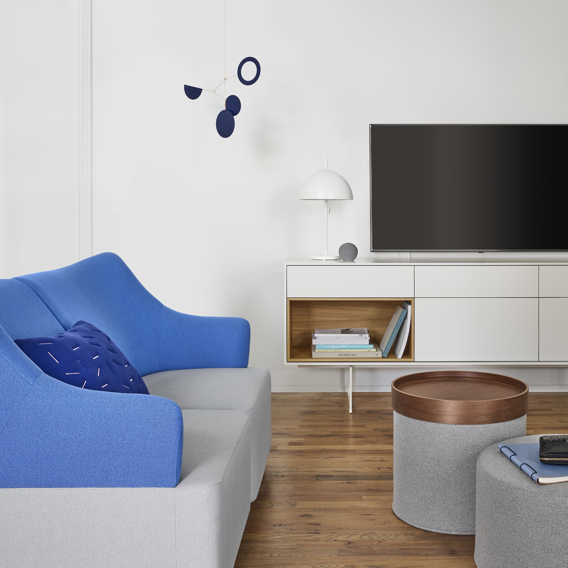 Plex Lounge Furniture in light grey and blue facing a light grey Drum Pouf and a light grey Drum Pouf with an oak tray. The scene is set in front of a white Aura Credenza with a television and various decorative elements.