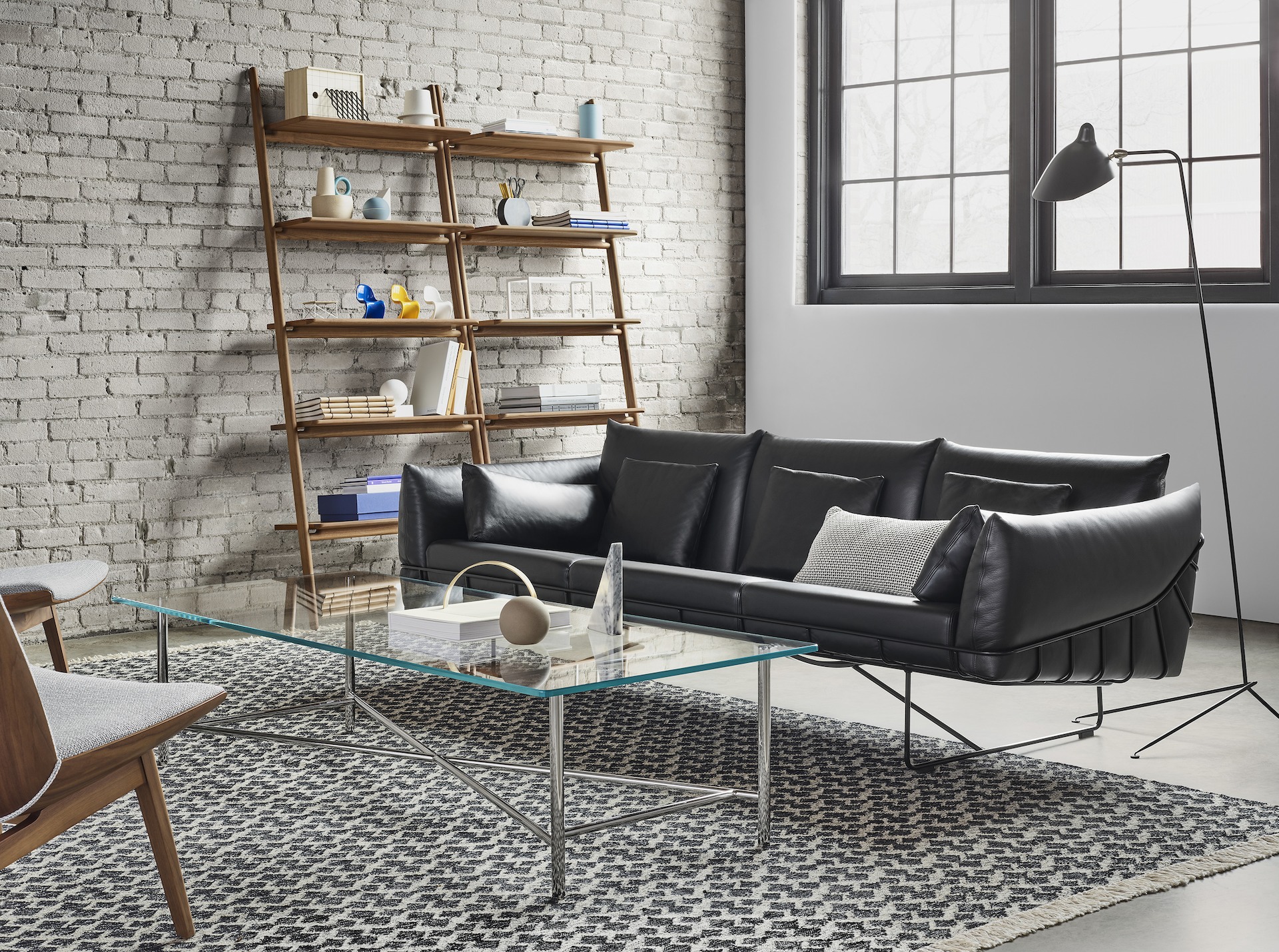 A black Wireframe Sofa and two Clamshell chairs around a glass and metal rectangular Layer Table. The scene is set on top of a Bowtie Kilim rug in front of a brick wall with a large window. Additional elements include two Folk Ladder Shelving units and a black Mantis BS1 B Floor Lamp. 
