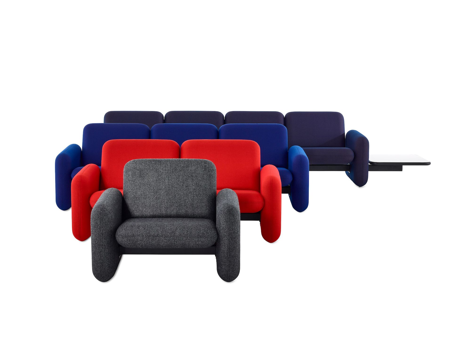 A grey Wilkes Modular Sofa Group Chair faces forward in font of a Wilkes Modular Sofa Group 2 Seat Sofa in red and a Wilkes Modular Sofa Group 3 Seat Sofa in blue and a Wilkes Modular Sofa Group 4 Seat Sofa and side table in navy blue.
