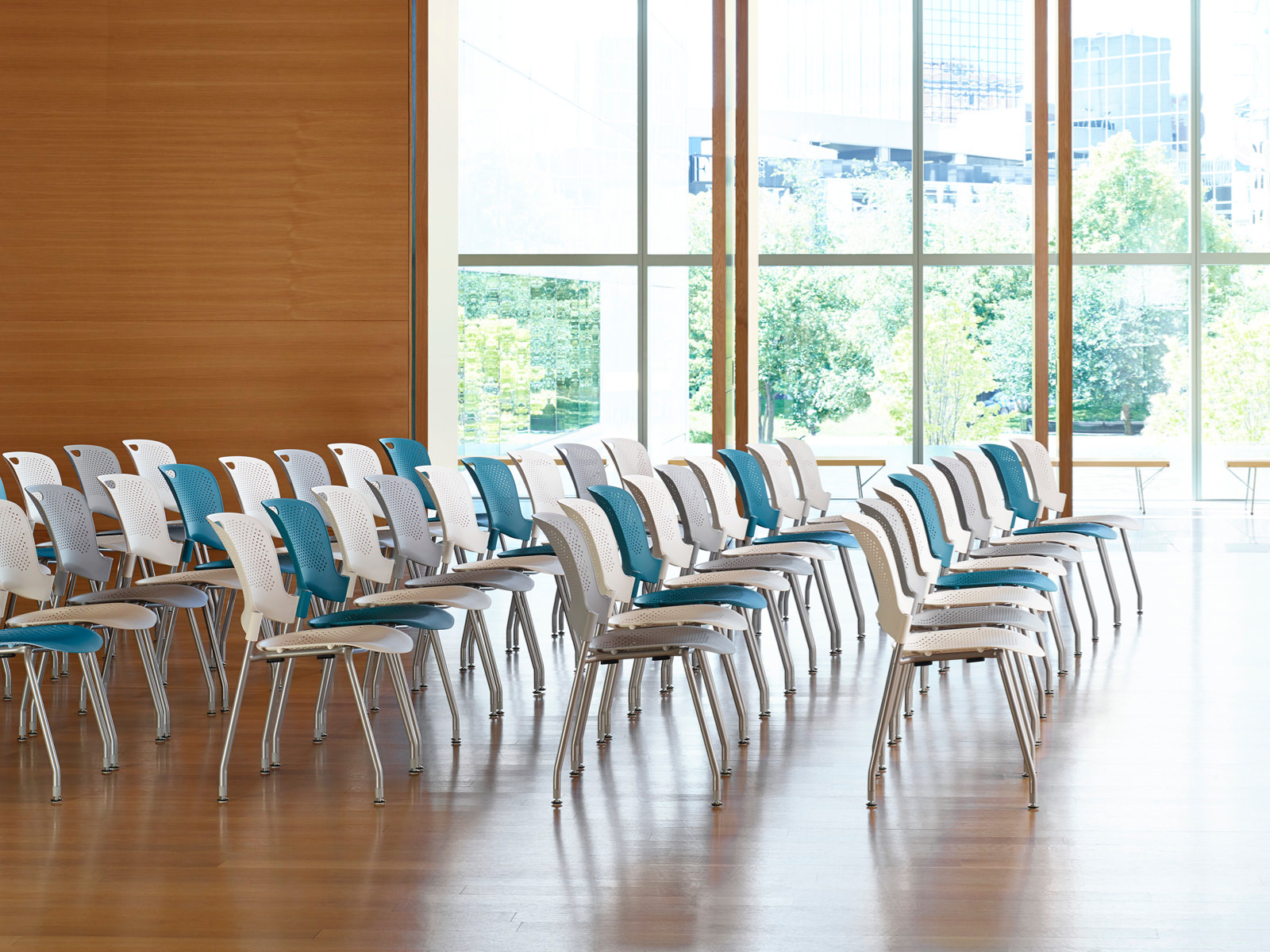 A presentation space with floor-to-ceiling windows contains five rows of Caper Stacking Chairs in white, light gray, and blue.