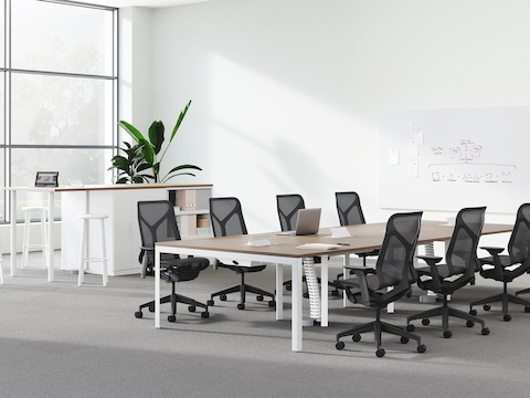 A rectangular table with wood top and white legs surrounded by 8 grey Cosm Chairs in front of a Layout Studio high desk.