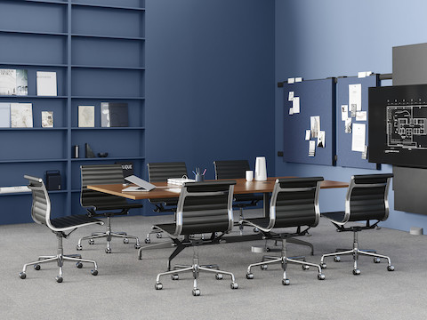 A conference table, surrounded by seven black Eames Aluminum Group chairs, is positioned in front of a large monitor. The table is powered by a Logic Reach wall start.