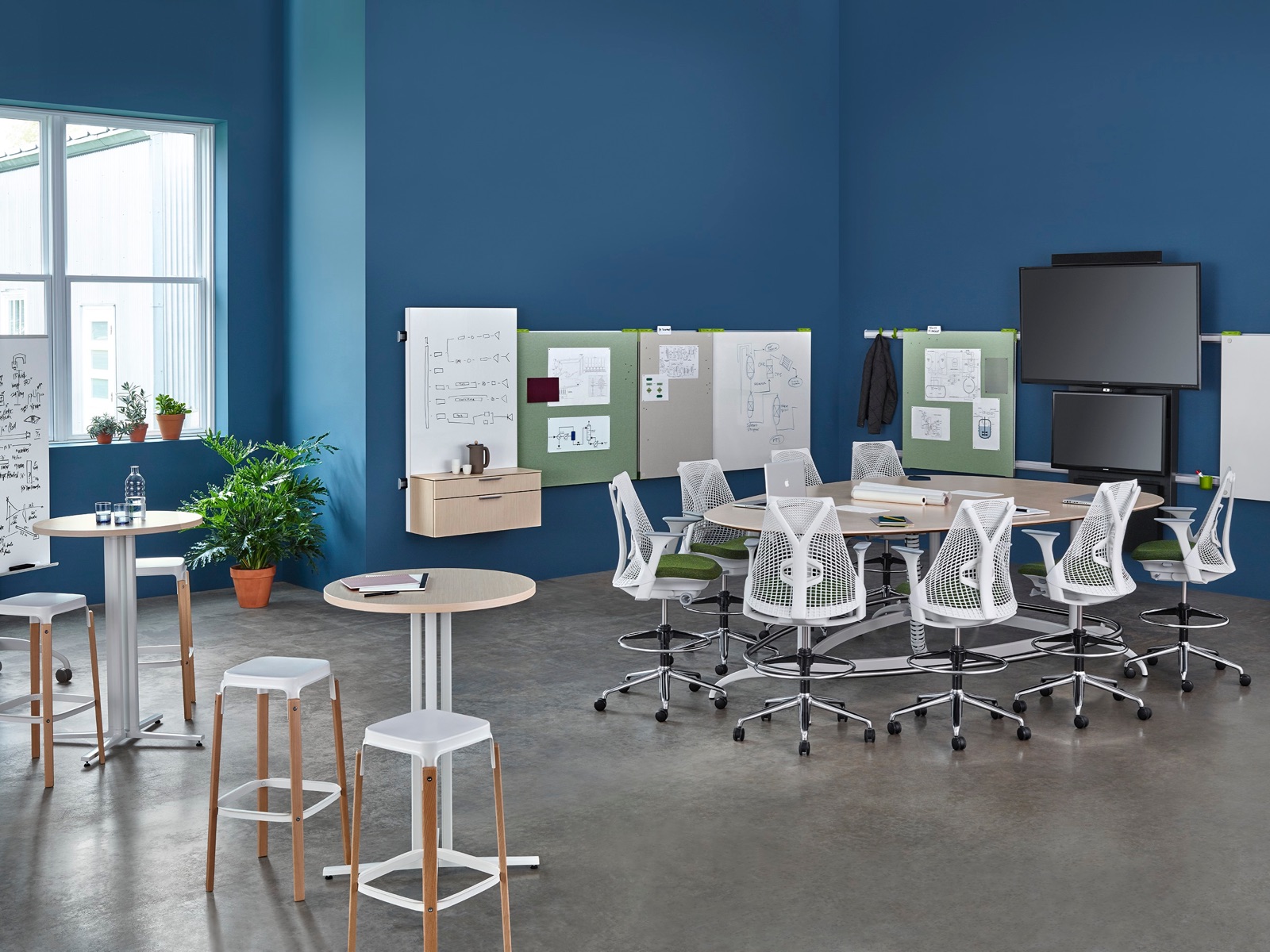 White Sayl Stools with green seat pads surround a teardrop Exclave table in a collaboration space with wall-hung display boards and dual monitors.