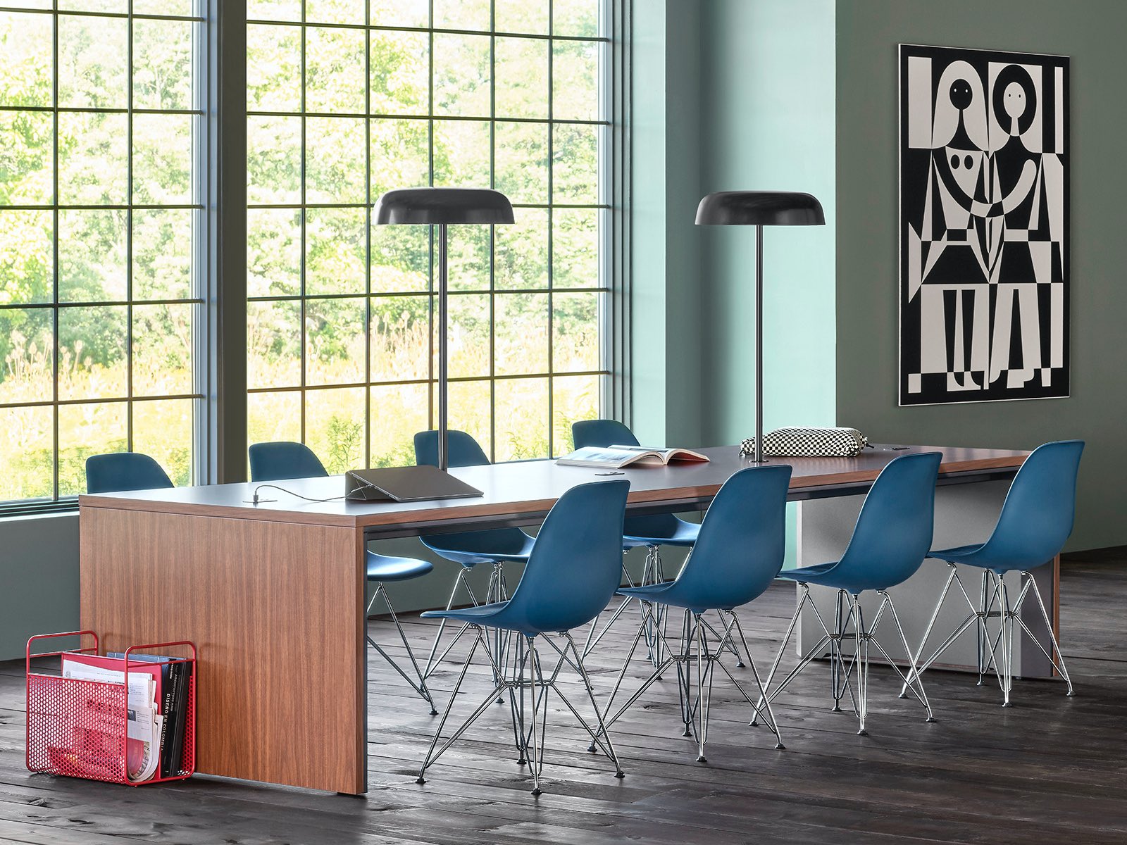 A community space with a Headway communal table surrounded by Eames Plastic side chairs and another nearby communal table behind a couch facing a TV.