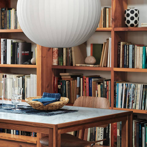 Ein Eames Molded Plywood Esszimmerstuhl mit einem Fußkreuz aus Metall neben einem rechteckigen Tisch mit Doppelrahmen und Bücherregalen im Hintergrund.