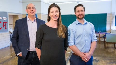 Two men and a woman stand smiling in a school classroom.