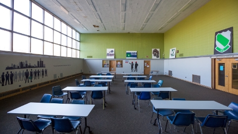 A classroom with blue chairs and tables with white tops, with illustrations on the walls and large, high windows. A classroom with blue chairs and tables with white tops, with illustrations on the walls and large, high windows.