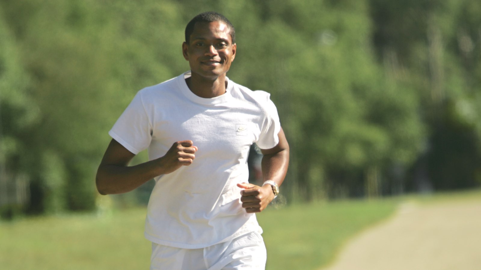 A man smiles while running in white t-shirt and shorts on a path outside near a forest.