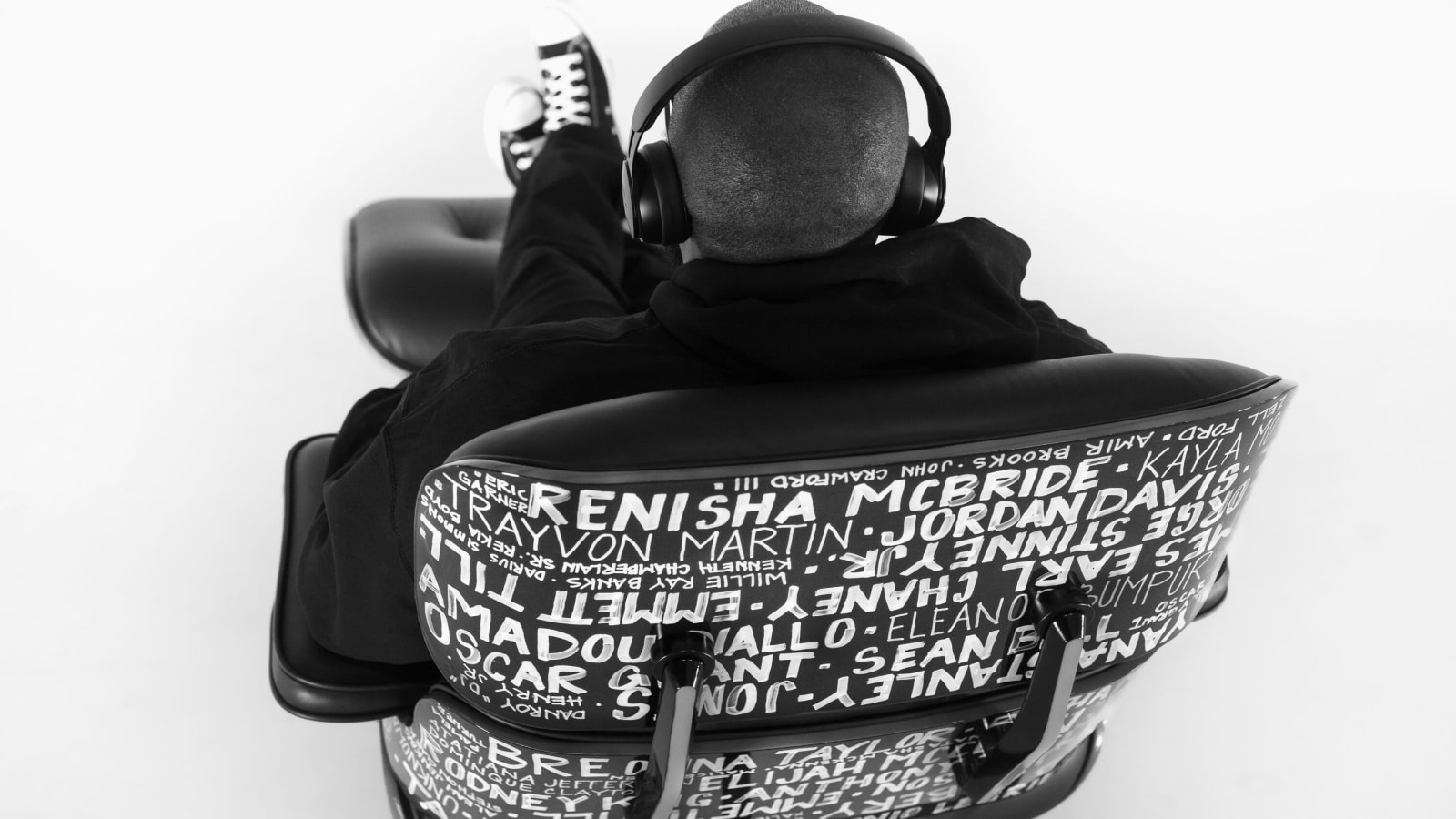 A man sitting in an Eames Lounge Chair and Ottoman with headphones on, viewed from behind. A man sitting in an Eames Lounge Chair and Ottoman with headphones on, viewed from behind.